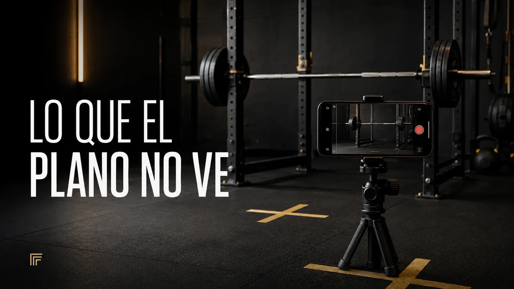 Un móvil sujeto en un soporte a media altura y nivelado graba de lado una barra cargada en un rack, en un gimnasio en penumbra; en el suelo hay dos marcas de cinta y en la pantalla del móvil la escena se ve más pequeña y desplazada.
